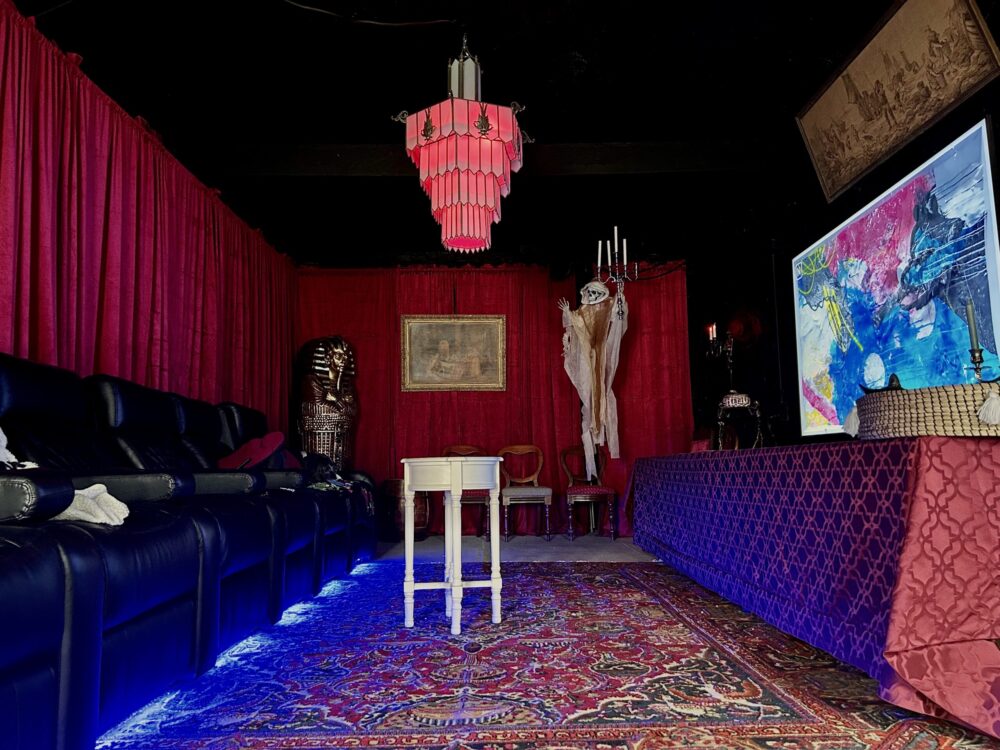 Gertrude's Cistern speakeasy lounge at 300 Clifton, with black leather reclining theater seats, red velvet drapes, a deco chandelier, and a skeleton on the wall.