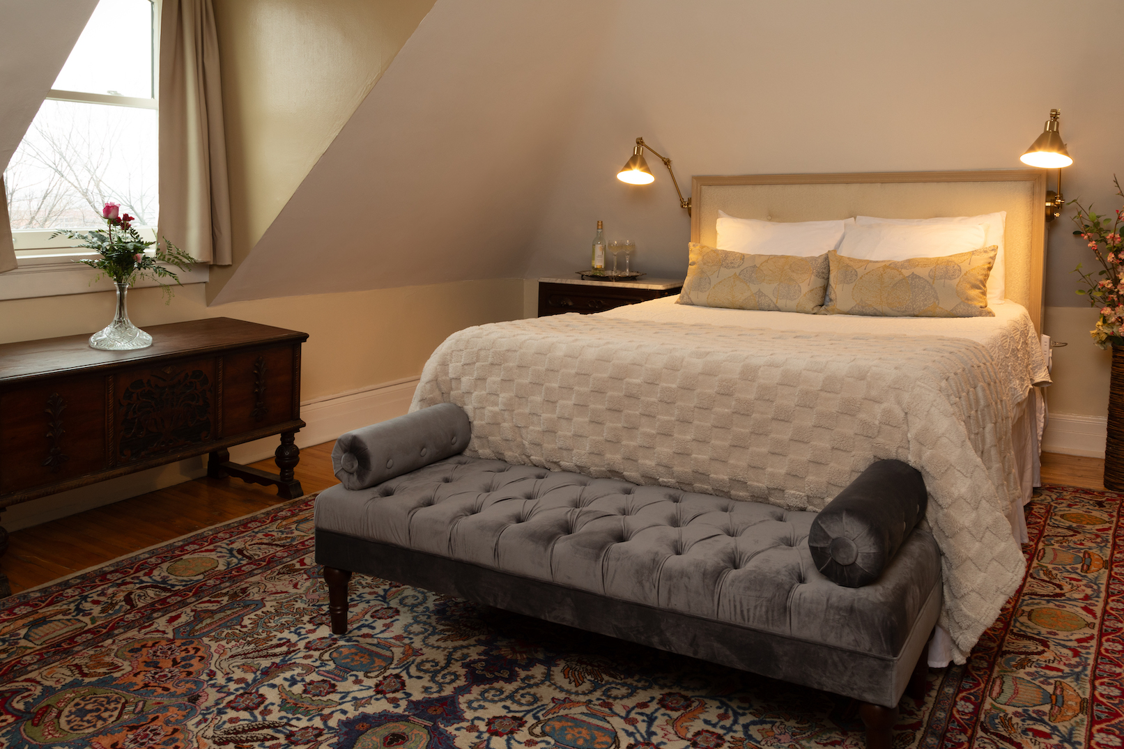 A dormer guest room at 300 Clifton with a cream upholstered bed, a grey tufted bench, a patterned rug and soft lamplight under a sloping ceiling.