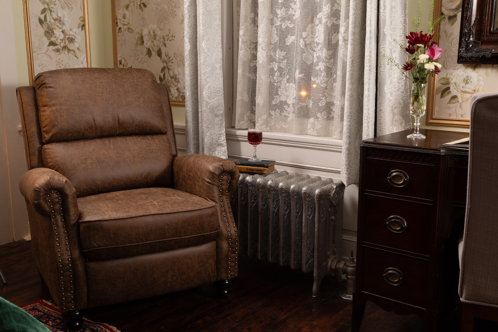 A leather armchair beside a lace-curtained window at 300 Clifton, a glass of red wine resting on a book atop the original cast-iron radiator