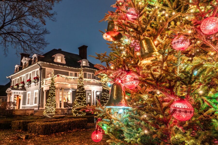 The 300 Clifton mansion glowing with warm holiday lights at night, framed by a Christmas tree hung with red and gold ornaments